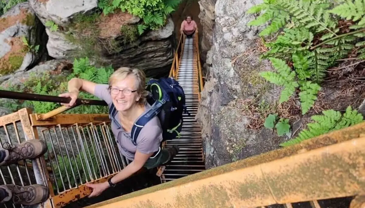 Zwei Personen steigen eine schmale Metalltreppe zwischen Felswänden hinauf; Farn und Wald im Hintergrund. | © DAV Markt Schwaben/Josephine Freikamp
