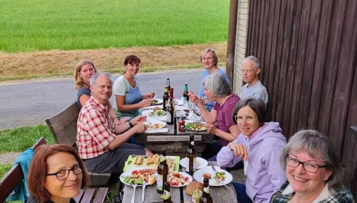 Elf Personen sitzen an Holztisch im Freien und essen gemeinsam; Wiese, Bäume und Hütte im Hintergrund. | © DAV Markt Schwaben/Foto Josephine Freikamp