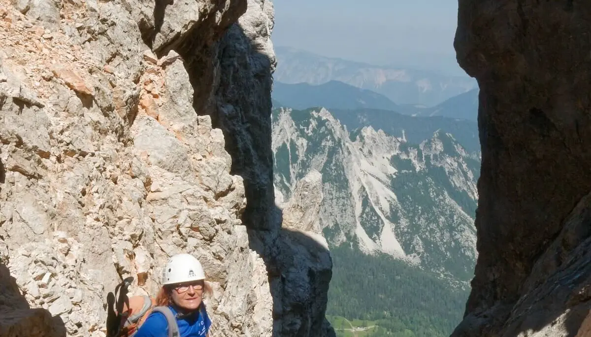 Person steht unter natürlichem Felsbogen mit Blick ins Tal; Kletterausrüstung und Bergkulisse im Hintergrund. | © DAV Markt Schwaben/Lutz Gründel