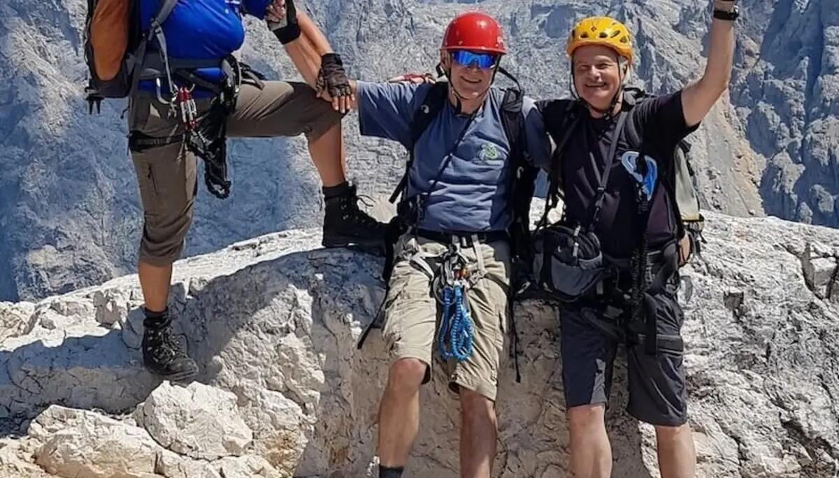 Drei Personen mit Kletterausrüstung stehen auf felsigem Gipfel; Bergpanorama und Wolken im Hintergrund. | © DAV Markt Schwaben/Helga Peters 