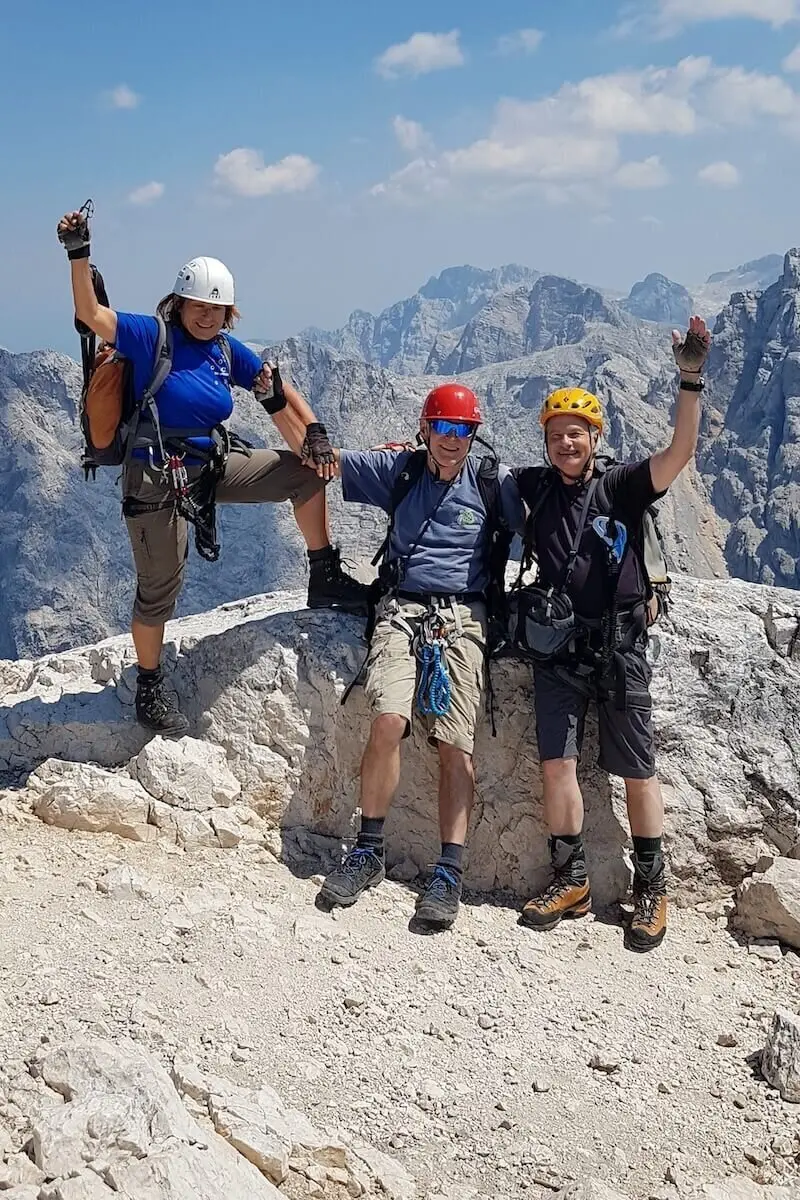 Drei Personen mit Kletterausrüstung stehen auf felsigem Gipfel; Bergpanorama und Wolken im Hintergrund. | © DAV Markt Schwaben/Helga Peters 
