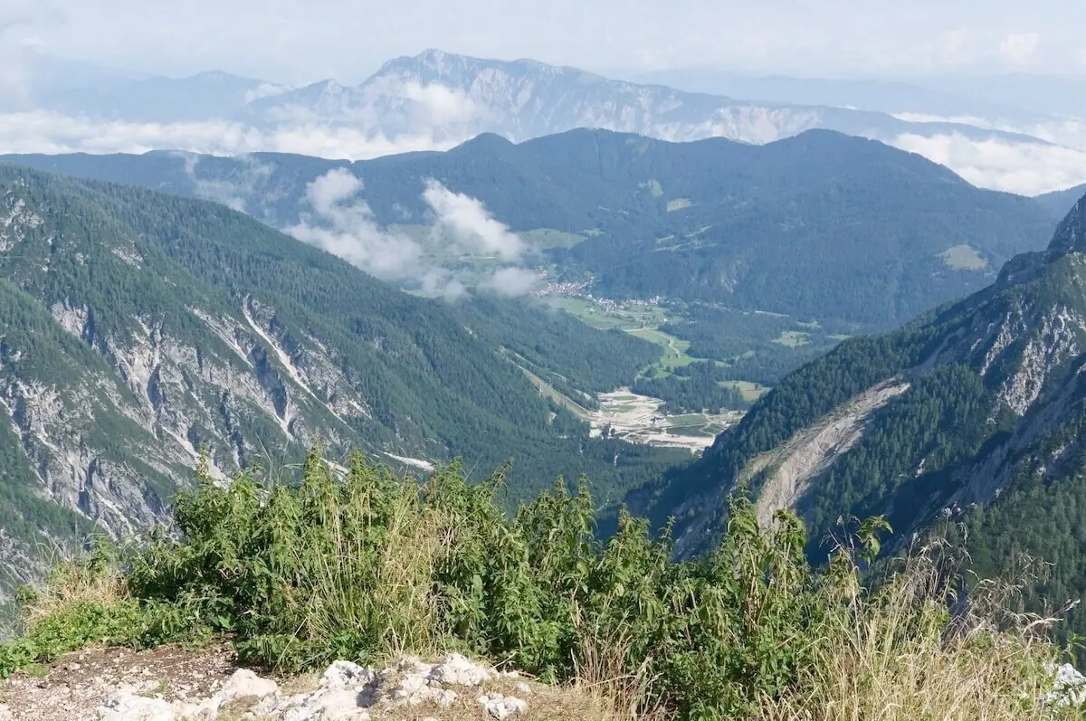 Hohe Gräser und Felsen im Vordergrund; dahinter grünes Tal mit Straße, Wald und Bergketten unter Wolken. | © DAV Markt Schwaben/Lutz Gründel