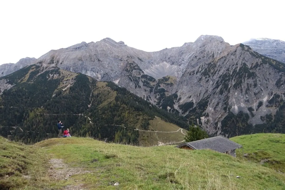 Zwei Wandernde stehen auf Wiese vor steilen Berggipfeln; Holzhütte rechts, Himmel bewölkt. | © DAV Markt Schwaben/Gerlinde Hübl
