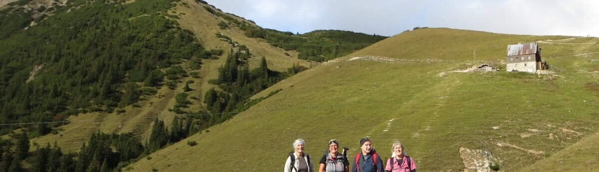 Vier Personen stehen auf Bergpfad vor Wiesen und Wald; Holzhütte rechts, Himmel leicht bewölkt. | © DAV Markt Schwaben/Gerlinde Hübl