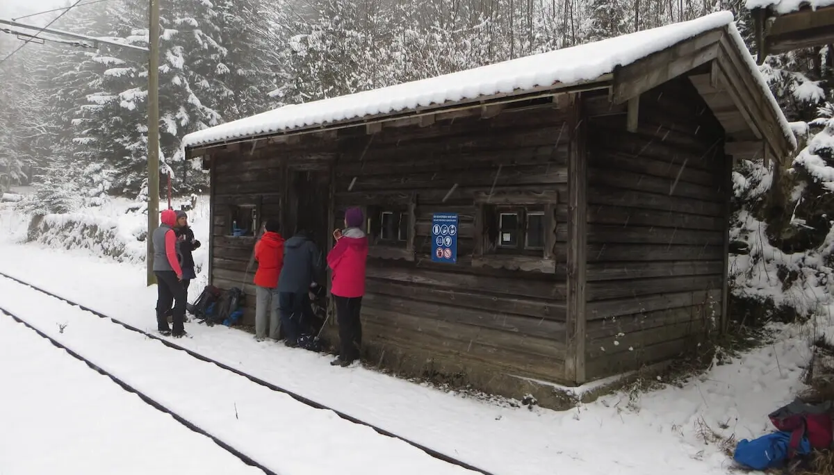 Mehrere Personen in Winterkleidung stehen vor einem Holzhaus mit Schildern; Schnee bedeckt Boden und Dach. | © DAV Markt Schwaben/Gerlinde Hübl