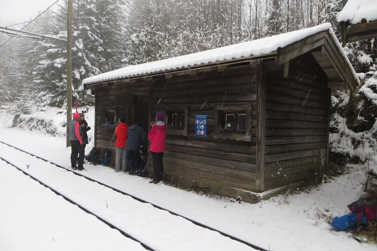 Mehrere Personen in Winterkleidung stehen vor einem Holzhaus mit Schildern; Schnee bedeckt Boden und Dach. | © DAV Markt Schwaben/Gerlinde Hübl