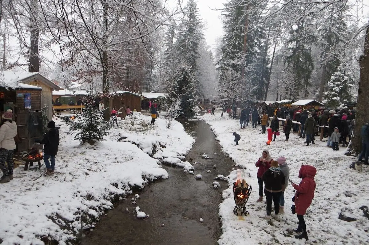 Menschen schlendern zwischen Holzständen im Schnee; Bach fließt durch die Mitte, Tannen im Hintergrund. | © DAV Markt Schwaben/Gerlinde Hübl