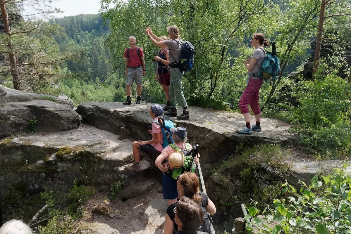 Acht Wandernde stehen auf Felsen im Wald; eine Person gestikuliert, Bäume und Vegetation umrahmen die Szene. | © DAV Markt Schwaben | Foto Lutz Gründel