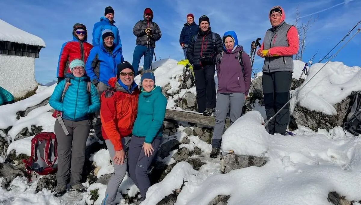 Dreizehn Personen posieren auf verschneitem Hang; Winterkleidung, Trekkingstöcke und Hütte im Hintergrund. | © DAV Markt Schwaben | Foto Markus Sellmeier