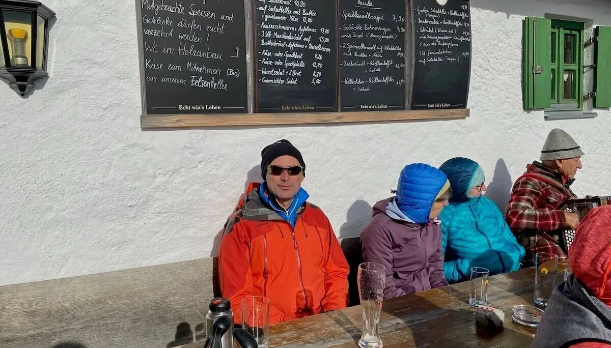 Gruppe sitzt vor rustikalem Gasthaus in der Sonne; Holztisch mit Gläsern, Speisekarte an weißer Wand. | © DAV Markt Schwaben | Foto Markus Sellmeier
