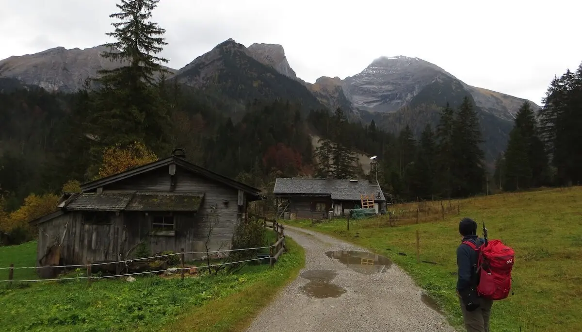 Person mit rotem Rucksack geht auf nassem Kiesweg zwischen zwei Holzhütten; Berge und Wald im Hintergrund. | © DAV Markt Schwaben | Foto Gerlinde Hübl