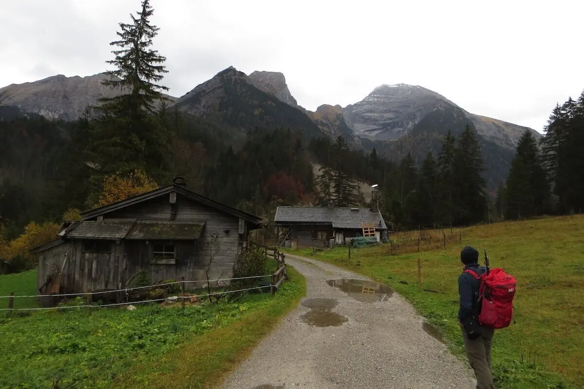 Person mit rotem Rucksack geht auf nassem Kiesweg zwischen zwei Holzhütten; Berge und Wald im Hintergrund. | © DAV Markt Schwaben | Foto Gerlinde Hübl