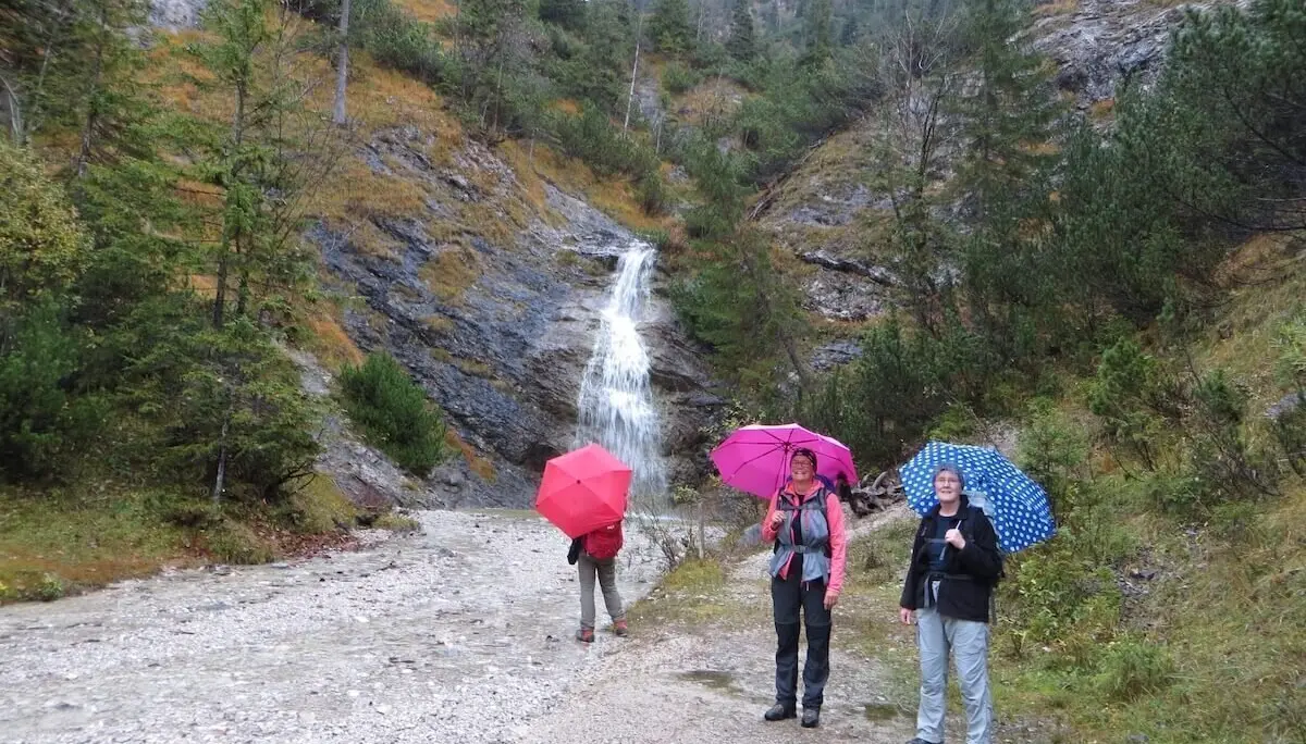 Drei Personen mit bunten Regenschirmen stehen vor kleinem Wasserfall; Wald und Felsen rahmen die Szene. | © DAV Markt Schwaben | Foto Gerlinde Hübl
