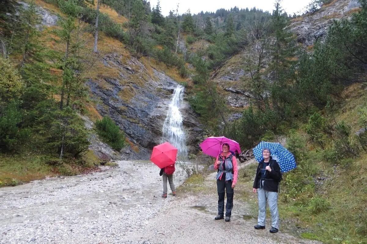 Drei Personen mit bunten Regenschirmen stehen vor kleinem Wasserfall; Wald und Felsen rahmen die Szene. | © DAV Markt Schwaben | Foto Gerlinde Hübl