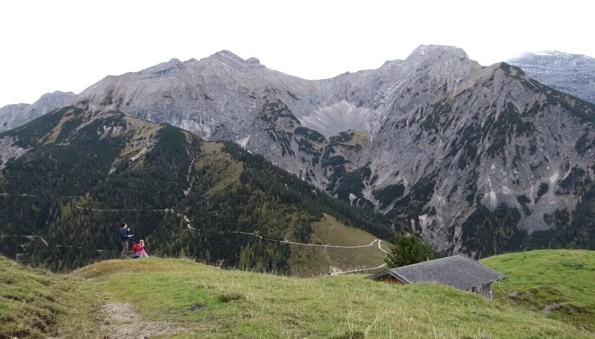 Zwei Wandernde stehen auf Wiese vor steilen Berggipfeln; Holzhütte rechts, Himmel bewölkt. | © DAV Markt Schwaben | Foto Gerlinde Hübl