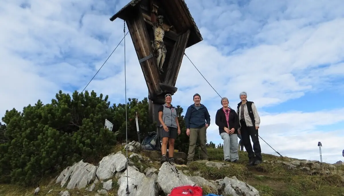 Vier Wandernde stehen am Gipfelkreuz des Kompar; Rucksäcke liegen im Vordergrund, Himmel teils bewölkt. | © DAV Markt Schwaben | Foto Gerlinde Hübl