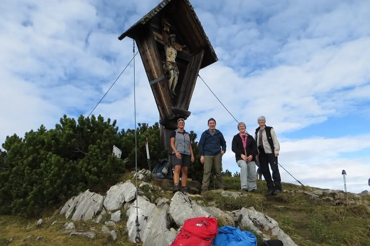 Vier Wandernde stehen am Gipfelkreuz des Kompar; Rucksäcke liegen im Vordergrund, Himmel teils bewölkt. | © DAV Markt Schwaben | Foto Gerlinde Hübl