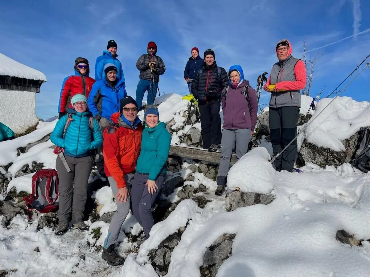 Dreizehn Personen posieren auf verschneitem Hang; Winterkleidung, Trekkingstöcke und Hütte im Hintergrund. | © DAV Markt Schwaben | Foto Markus Sellmeier