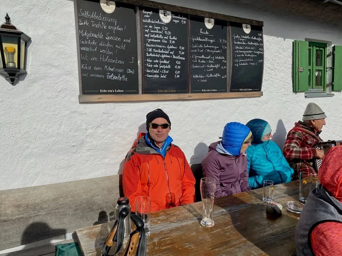 Gruppe sitzt vor rustikalem Gasthaus in der Sonne; Holztisch mit Gläsern, Speisekarte an weißer Wand. | © DAV Markt Schwaben | Foto Markus Sellmeier