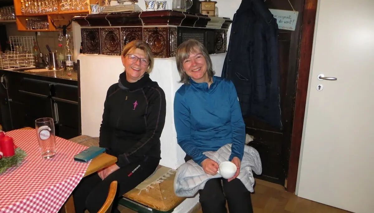 Zwei Personen sitzen entspannt auf Holzbank; hinter ihnen Ofen mit Deko, Tisch mit Bier und Adventsgesteck. | © DAV Markt Schwaben | Foto Gerlinde Hübl