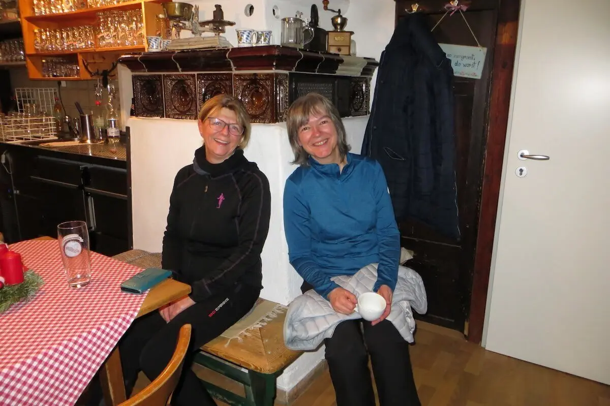 Zwei Personen sitzen entspannt auf Holzbank; hinter ihnen Ofen mit Deko, Tisch mit Bier und Adventsgesteck. | © DAV Markt Schwaben | Foto Gerlinde Hübl