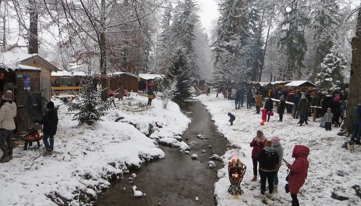 Menschen schlendern zwischen Holzständen im Schnee; Bach fließt durch die Mitte, Tannen im Hintergrund. | © DAV Markt Schwaben | Foto Gerlinde Hübl