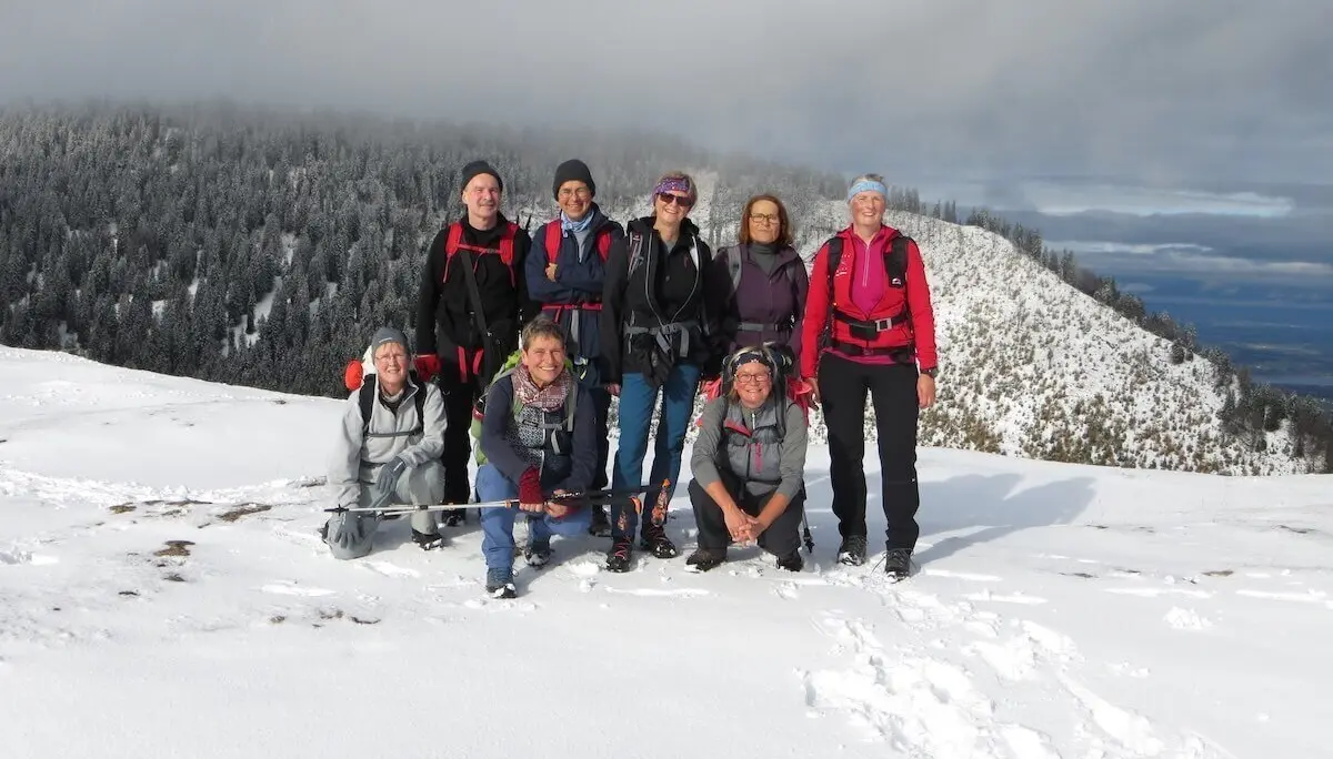 Acht Personen posieren auf verschneitem Gipfel; vier stehen, vier knien, Bäume und Wolken im Hintergrund. | © DAV Markt Schwaben | Foto Gerlinde Hübl