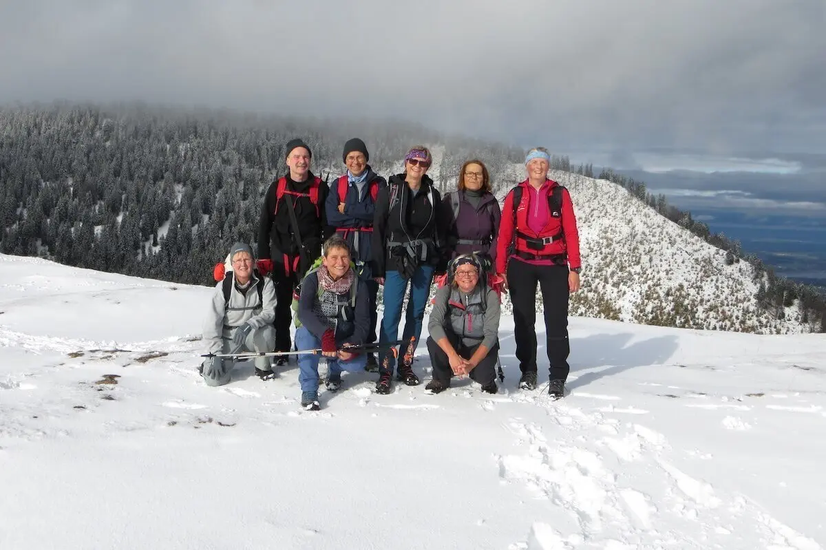 Acht Personen posieren auf verschneitem Gipfel; vier stehen, vier knien, Bäume und Wolken im Hintergrund. | © DAV Markt Schwaben | Foto Gerlinde Hübl
