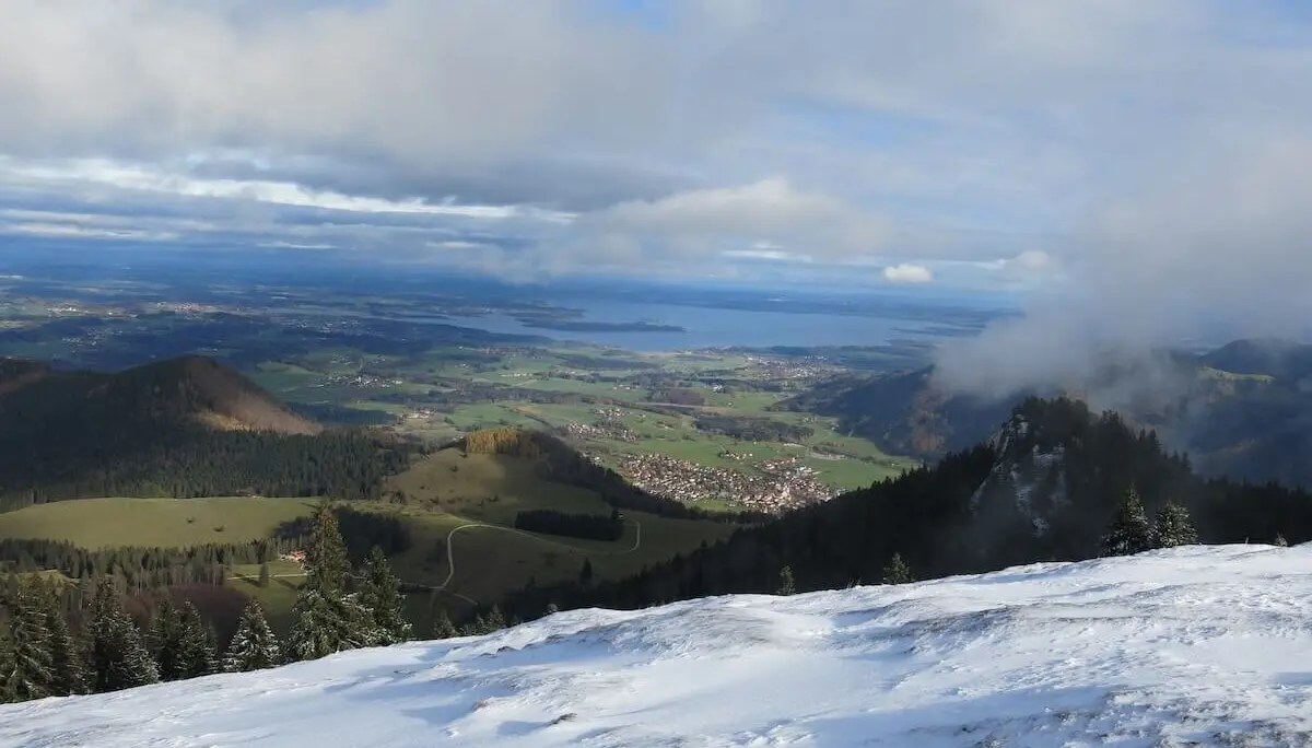 Schneebedeckter Hang mit Sicht auf grüne Täler und Chiemsee; Wolken und blauer Himmel im Hintergrund. | © DAV Markt Schwaben | Foto Erwin Matzinger