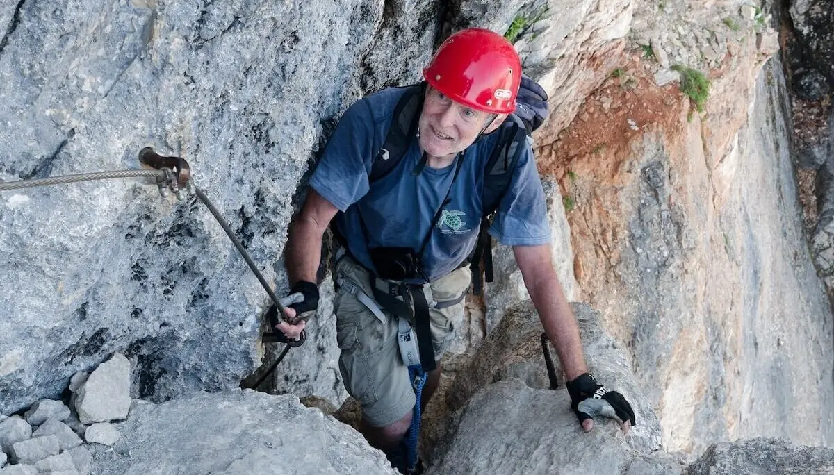 Kletterer mit rotem Helm und Handschuhen steigt durch steile Felsrinne; graue und rötliche Gesteinsstruktur. | © DAV Markt Schwaben | Foto Lutz Gründel