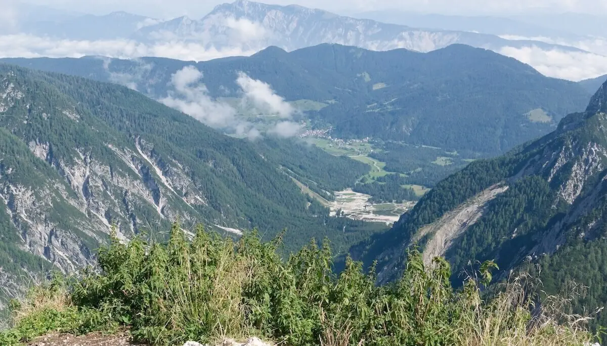 Hohe Gräser und Felsen im Vordergrund; dahinter grünes Tal mit Straße, Wald und Bergketten unter Wolken. | © DAV Markt Schwaben | Foto Lutz Gründel