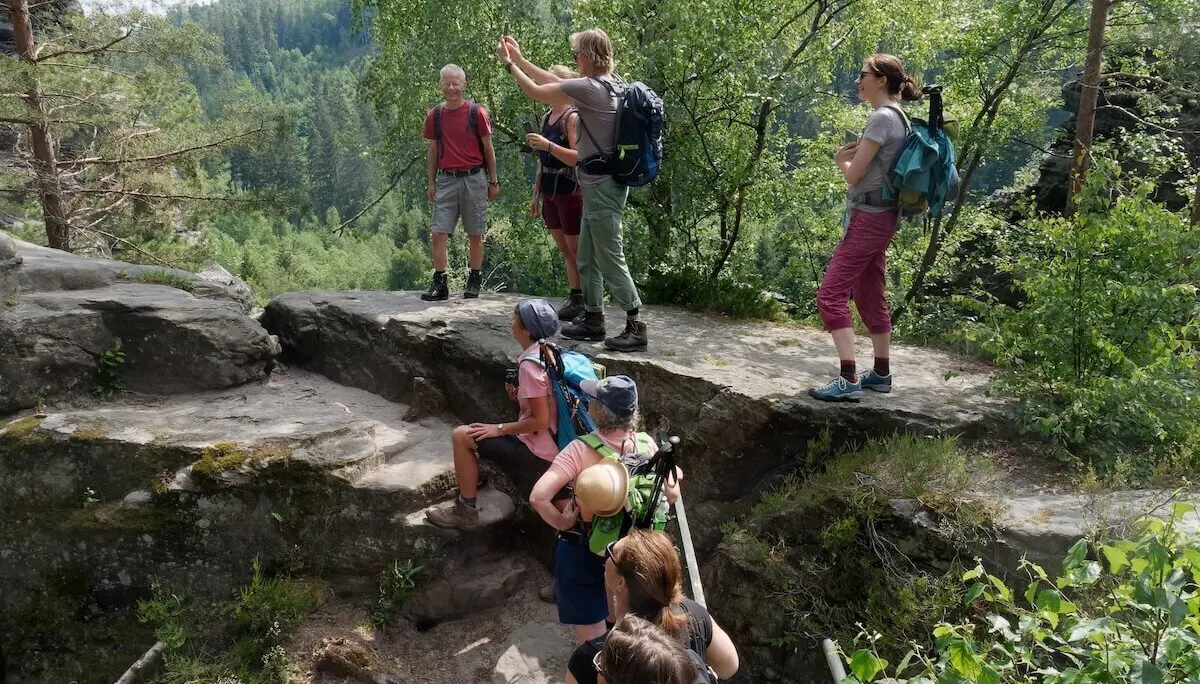 Acht Wandernde stehen auf Felsen im Wald; eine Person gestikuliert, Bäume und Vegetation umrahmen die Szene. | © DAV Markt Schwaben | Foto Lutz Gründel