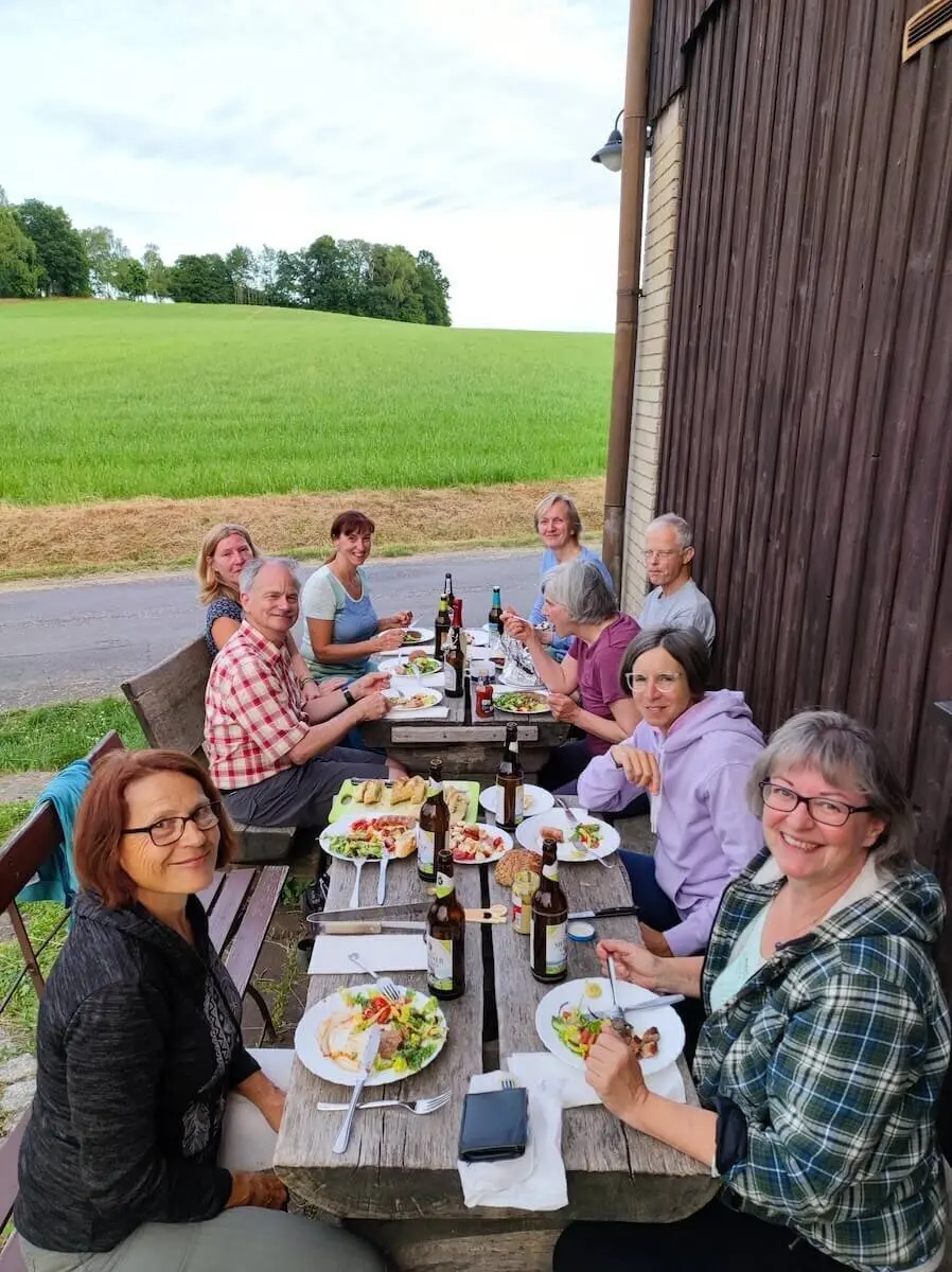 Elf Personen sitzen an Holztisch im Freien und essen gemeinsam; Wiese, Bäume und Hütte im Hintergrund. | © DAV Markt Schwaben | Foto Josephine Freikamp