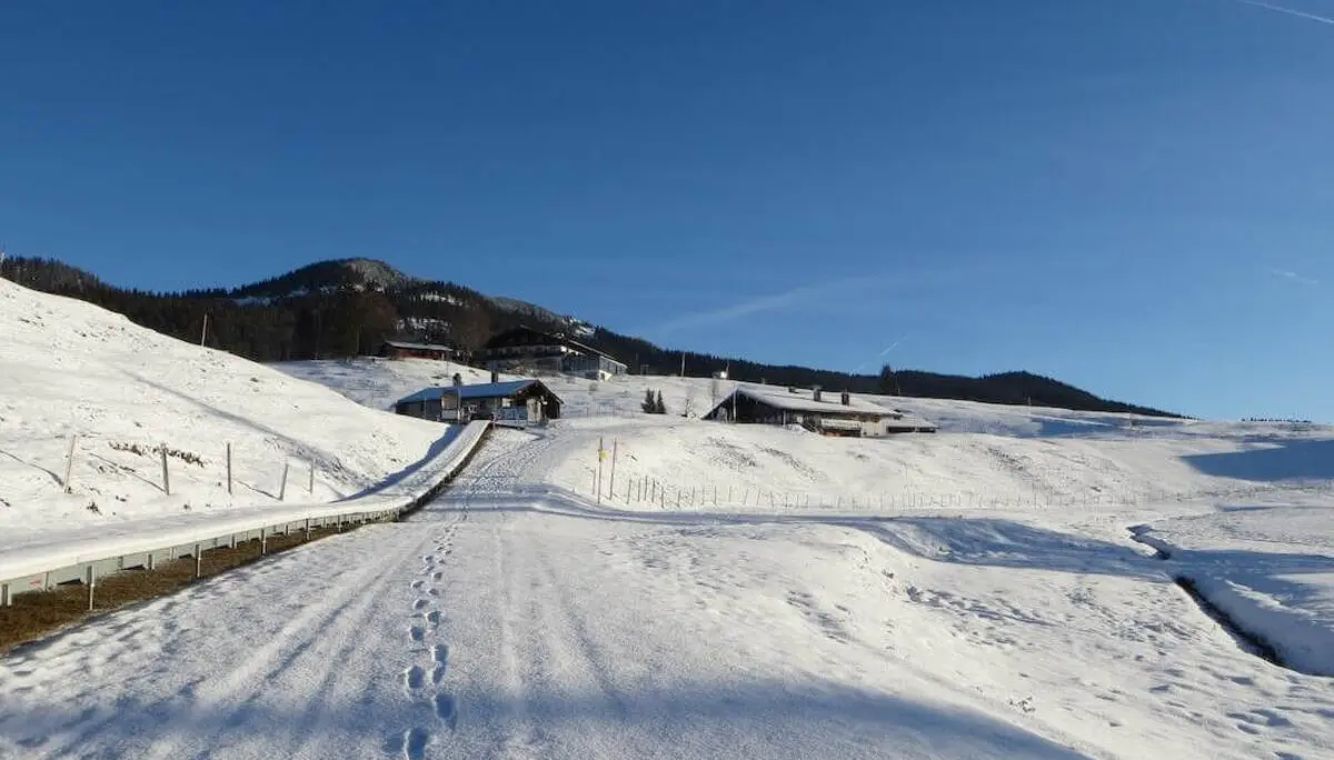 Verschneiter Fahrweg mit Fußspuren führt über die Alm zu mehreren Gebäuden; dahinter liegen sanfte, schneebedeckte Hügel. | © DAV Markt Schwaben / Gerlinde Hübl
