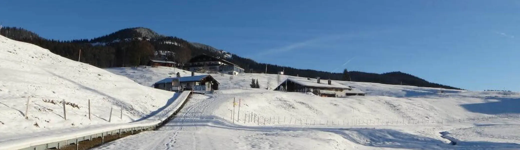 Verschneiter Fahrweg mit Fußspuren führt über die Alm zu mehreren Gebäuden; dahinter liegen sanfte, schneebedeckte Hügel. | © DAV Markt Schwaben / Gerlinde Hübl