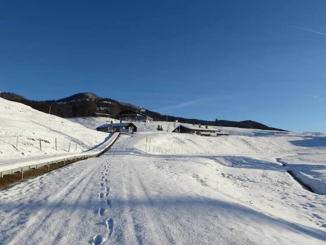 Verschneiter Fahrweg mit Fußspuren führt über die Alm zu mehreren Gebäuden; dahinter liegen sanfte, schneebedeckte Hügel. | © DAV Markt Schwaben / Gerlinde Hübl