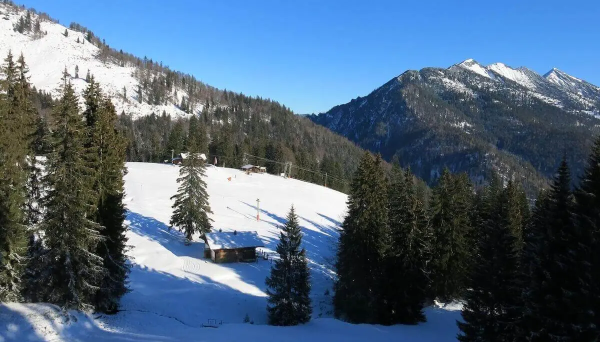 Zwischen bewaldeten Berghängen liegt eine schneebedeckte Alm mit mehreren Hütten; hohe Gipfel ragen im Hintergrund auf. | © DAV Markt Schwaben / Erwin Matzinger