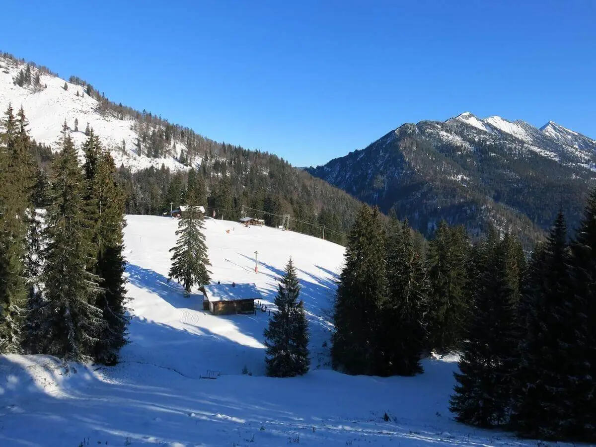 Zwischen bewaldeten Berghängen liegt eine schneebedeckte Alm mit mehreren Hütten; hohe Gipfel ragen im Hintergrund auf. | © DAV Markt Schwaben / Erwin Matzinger