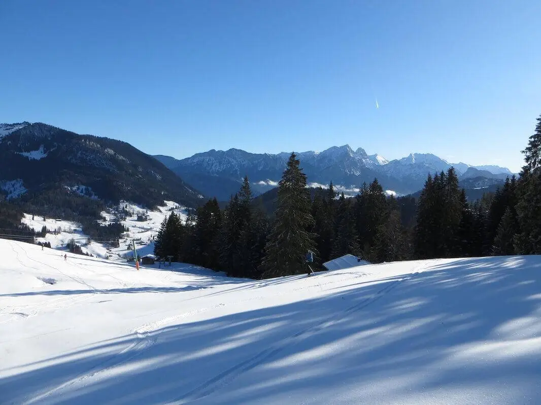 Weite, verschneite Hänge führen zu dunklen Waldflächen; dahinter erstreckt sich eine Gebirgskette unter klarem Himmel. | © DAV Markt Schwaben / Gerlinde Hübl