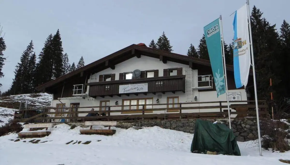Die Traunsteiner Hütte steht am Waldrand oberhalb einer schneebedeckten Fläche; vor dem Gebäude wehen mehrere Fahnen. | © DAV Markt Schwaben / Gerlinde Hübl