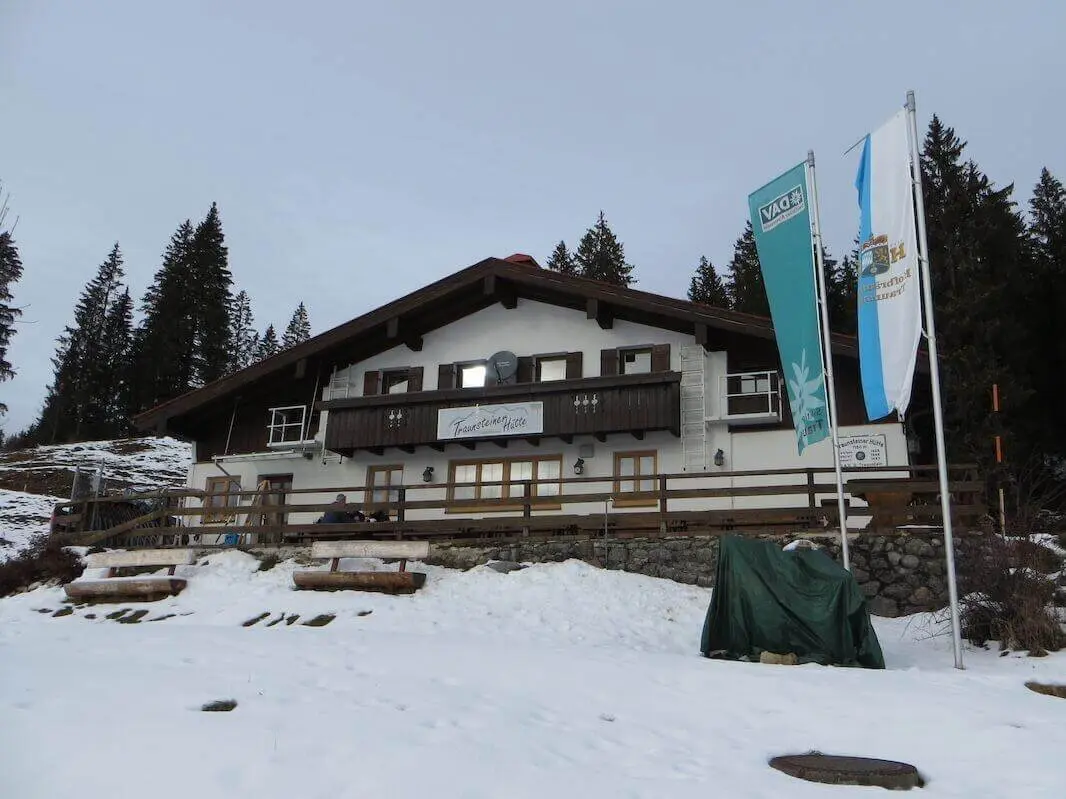 Die Traunsteiner Hütte steht am Waldrand oberhalb einer schneebedeckten Fläche; vor dem Gebäude wehen mehrere Fahnen. | © DAV Markt Schwaben / Gerlinde Hübl