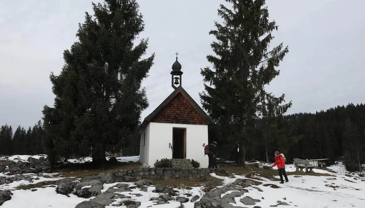 Kleine weiße Kapelle mit Dachreiter steht zwischen hohen Tannen auf einer teils schneebedeckten Almfläche mit Felsen. | © DAV Markt Schwaben / Erwin Matzinger