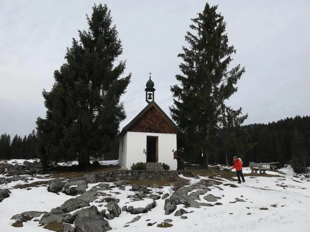 Kleine weiße Kapelle mit Dachreiter steht zwischen hohen Tannen auf einer teils schneebedeckten Almfläche mit Felsen. | © DAV Markt Schwaben / Erwin Matzinger