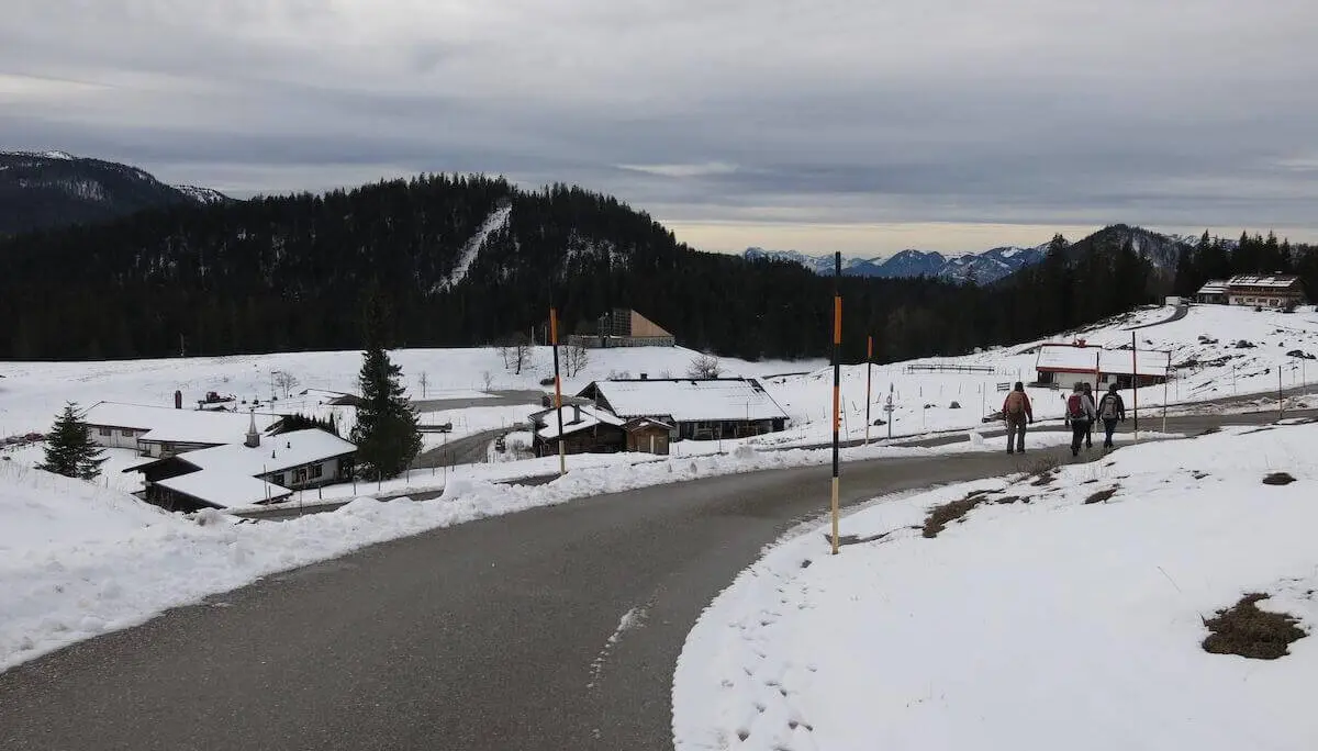 Eine geräumte Straße führt durch die verschneite Almlandschaft; mehrere Wandernde gehen an Gebäuden vorbei Richtung Tal. | © DAV Markt Schwaben / Erwin Matzinger