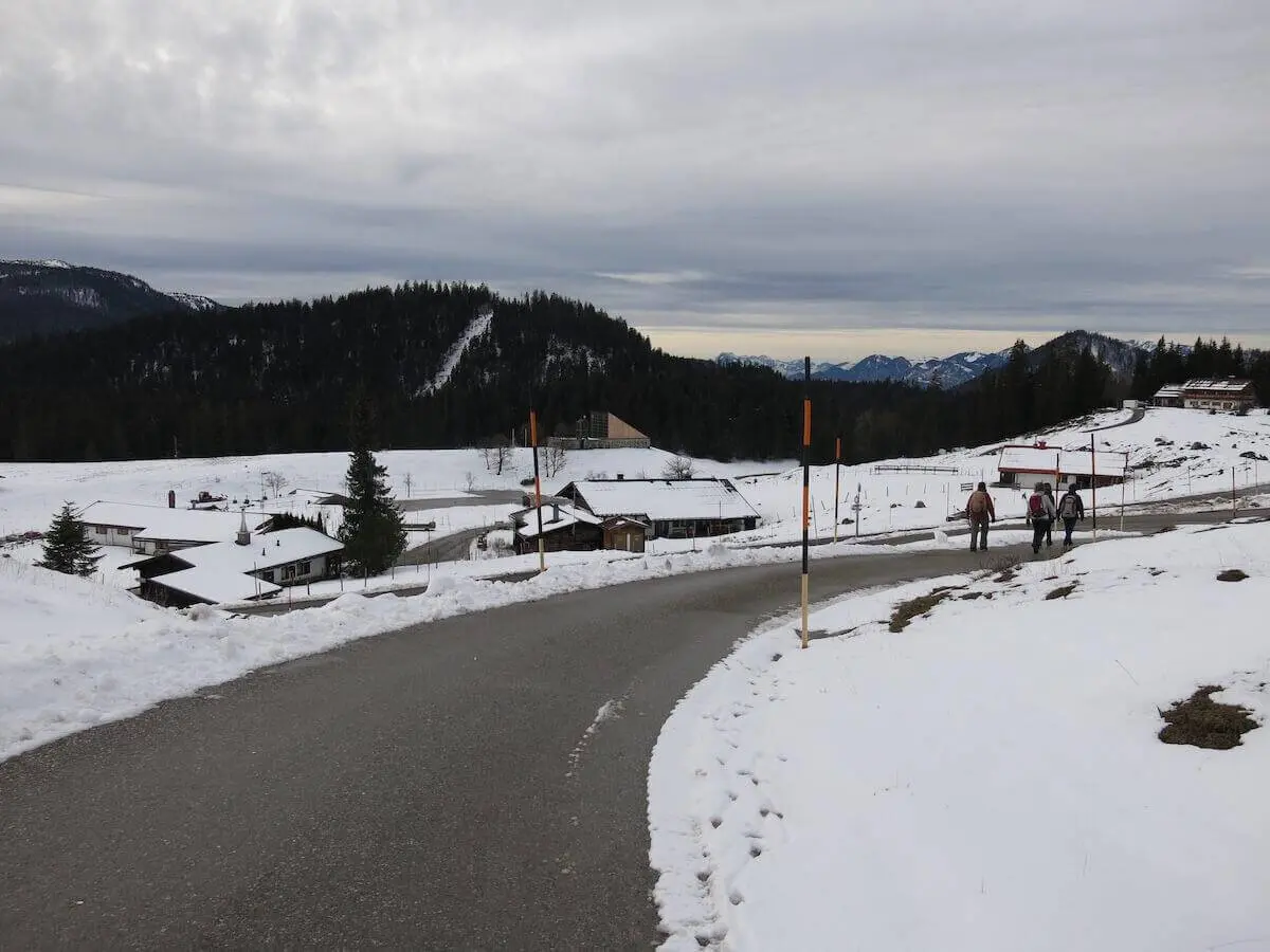 Eine geräumte Straße führt durch die verschneite Almlandschaft; mehrere Wandernde gehen an Gebäuden vorbei Richtung Tal. | © DAV Markt Schwaben / Erwin Matzinger