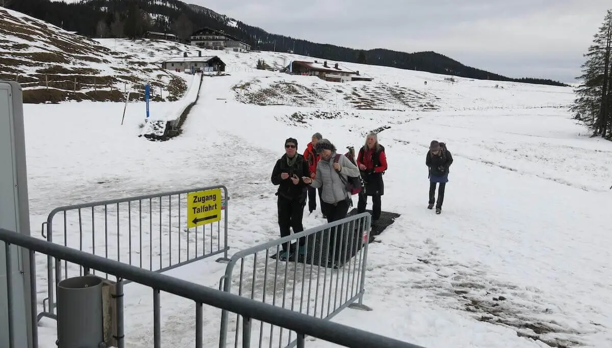Mehrere Wandernde erreichen über verschneiten Boden den Zugang zur Gondel-Talfahrt; Almhäuser stehen im Hintergrund. | © DAV Markt Schwaben / Erwin Matzinger