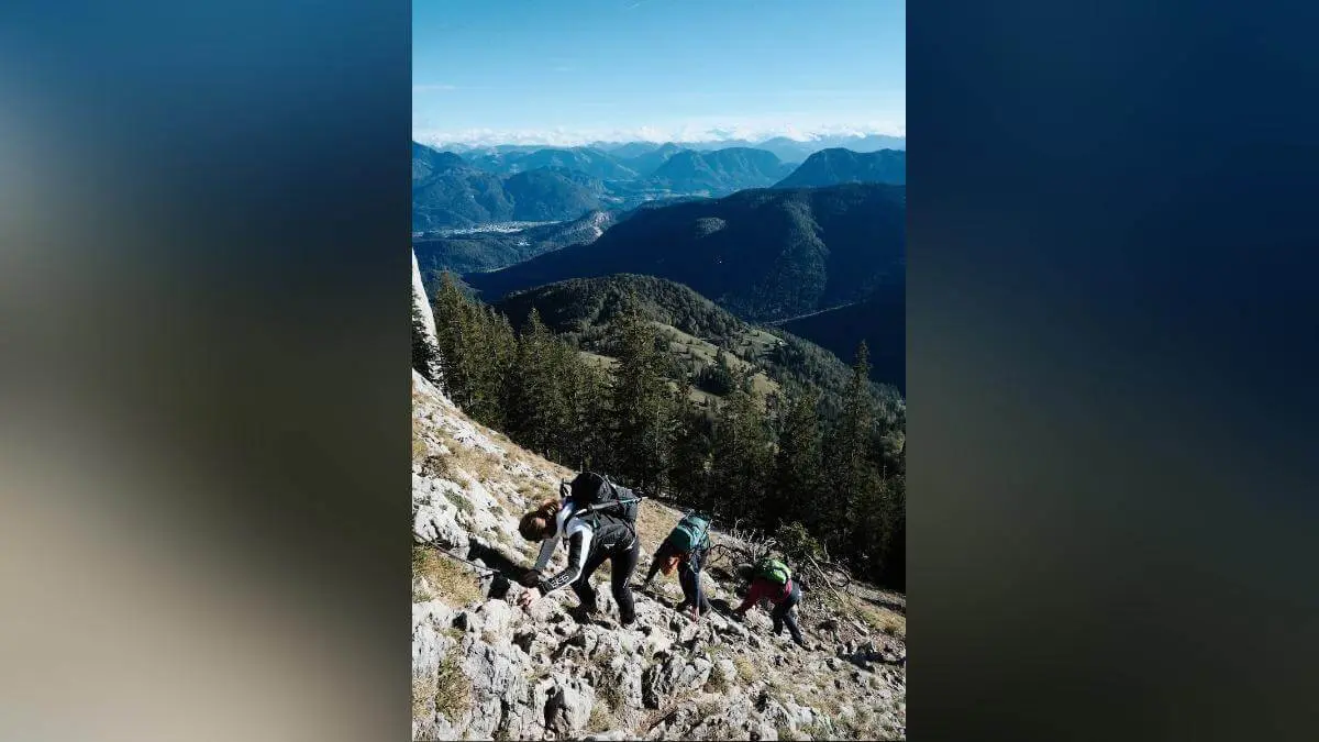 Drei Wandernde steigen über steile Felsen auf, mit Rucksäcken und weitem Blick auf bewaldete Berge unter blauem Himmel. | © DAV Markt Schwaben/Bernhard Zech