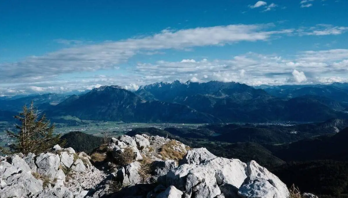 Blick von einem felsigen Hang über Täler, Felder und ferne Alpenberge unter teils bewölktem Himmel. | © DAV Markt Schwaben/Bernhard Zech