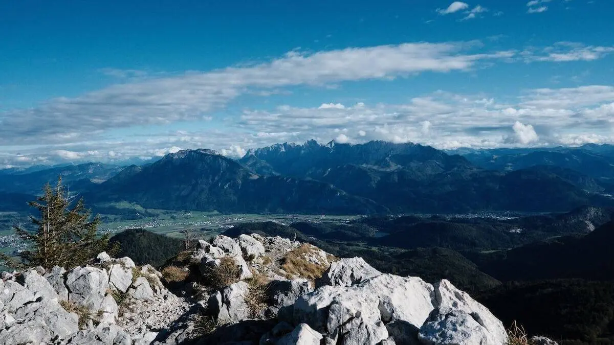 Blick von einem felsigen Hang über Täler, Felder und ferne Alpenberge unter teils bewölktem Himmel. | © DAV Markt Schwaben/Bernhard Zech