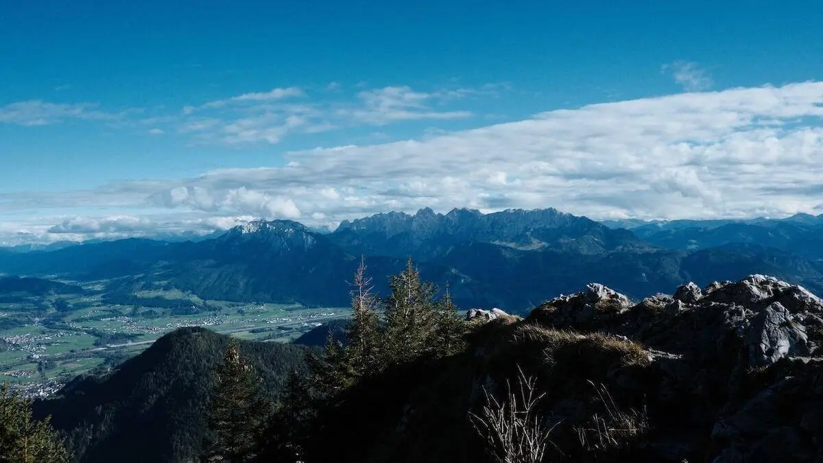 Panorama mit Felsen, Nadelbäumen und weitem Blick über grüne Täler bis zu wolkenverhangenen Alpenbergen unter blauem Himmel. | © DAV Markt Schwaben/Bernhard Zech
